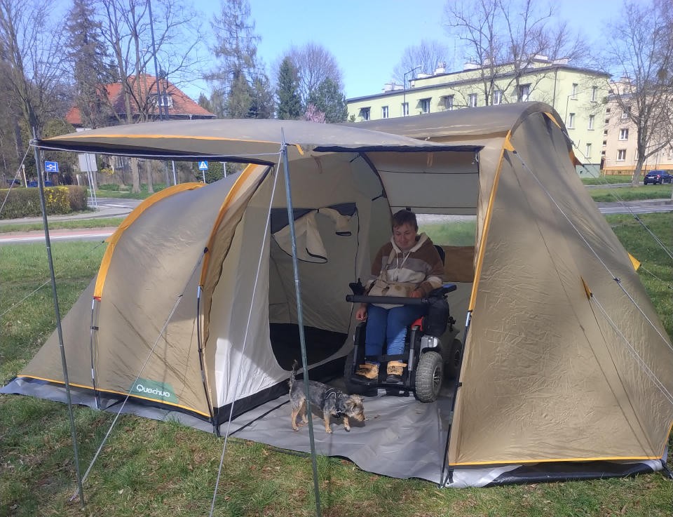 W przedsionku kempingowe go namiotu, na srebrzystej plandece, stoi elektryczny wózek z pasażerką. Przed nią mały kudłaty pies ogląda namiot od środka.