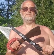 Zdjęcie pokazuje mężczyznę w średnim wieku bez koszulki, w szortach i okularach słonecznych, stojącego na tle obozowiska. Przed sobą trzyma zardzewiały tasak do gałęzi, skrzyżowany z ręczną piłą do drewna.